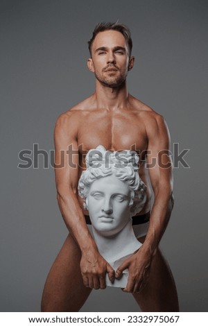 An alluring male model with a perfect physique confidently poses in nothing but underwear, juxtaposed against a bust of an ancient Greek sculpture on a gray background