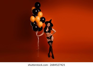 Alluring witch posing confidently with balloons against vibrant orange backdrop embracing Halloween magic and playful autumn vibes