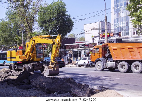Almaty, Kazakhstan - Sep 6, 2017:\
Reconstruction of street. Yellow excavator & orange truck\
against cafe &\
buildings