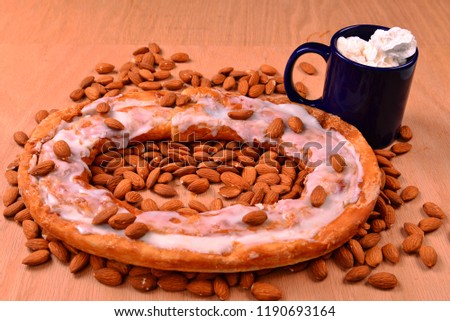 Almond Kringle on a bed of almonds laid over an red oak surface with a cup of hot chocolate. 