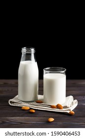 Almond milk in a glass mug and a refillable glass bottle, with almond nuts on a wooden table and black background.