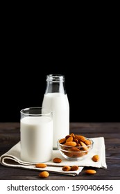 Almond milk in a glass mug and a refillable glass bottle, with almond nuts on a wooden table and black background.