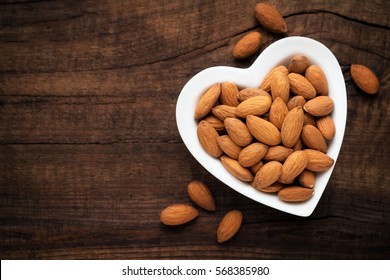 Almonds In A White Heart Shaped Bowl Against Dark Rustic Wooden Background. Overhead View