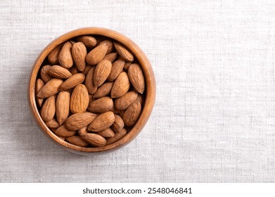 Almonds in a wooden bowl on linen fabric, with empty space for text. Shelled, raw, whole and dried nuts, seeds and fruits of Prunus dulcis. Ready-to-eat as snack, or used as an ingredient for baking.