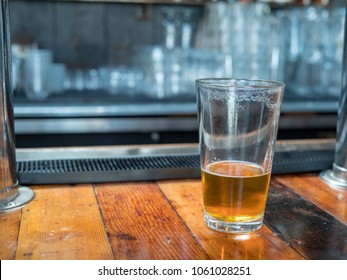 Almost Empty Pint Glass Of Beer Sitting On Woodencountertop For Last Call. Bar In Background