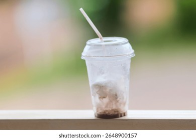 An almost empty plastic cup with a lid and a straw stands on a white ledge against a blurred green background.