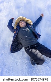 Almost Full Body Portrait Of Asian Fat Man Wearing Eyeglasses In Blue Overcoat With T-shirt Inside And Blue Jeans Lays Down On The Snow Field. He Look Funny.