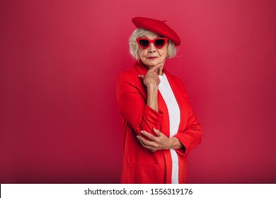 Almost full size picture of stylish fasion icon. Old woman in red coat and beret posing alone. Looking straight through sunglasses. Hold hand under chin. Isolated over red background