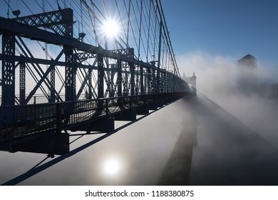 An almost silhouette of a beautiful bridge stands over a thick morning fog covering the river water below as the suns reflection tries to shine through but succeeding up above in the blue sky.