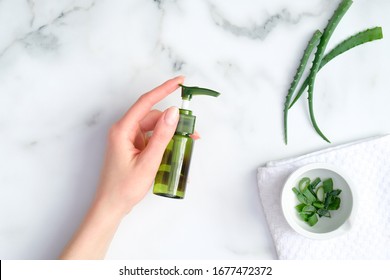 Aloe Vera Gel Green Plastic Bottle In Female Hand On Marble Background With Aloe Plant Stems. Natural Organic Lotion For Hand And Face Skin Treatment. Top View, Flat Lay