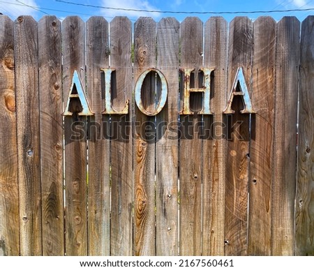 Aloha sign made of light blue rusted metal letters on a weathered wooden fence in the sun. A sliver of blue sky with clouds in the upper part of frame.