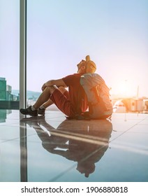 Alone Backpacker Traveler Sitting On The Airport Terminal Floor Near The Window, Looking At The Hall Ceiling And Waiting For Boarding At Aircraft Which Prepared For Flight Departure.