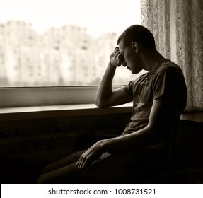 Alone broken sullen young teen guy person sit waiting look on light white gloomy city copyspace in window. Male addiction psychological emotion portrait and space for text on dark home room background