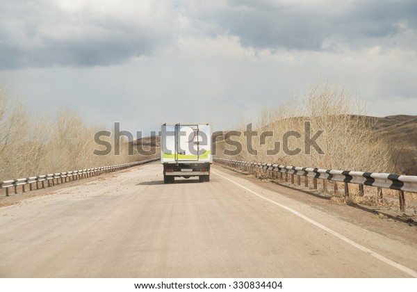 Alone clean Truck driving on clear spring\
asphalt road against blue sky with white clouds, empty mountain\
horizon with dry trees and forest along highway Back view car Idea\
symbol of logistics
