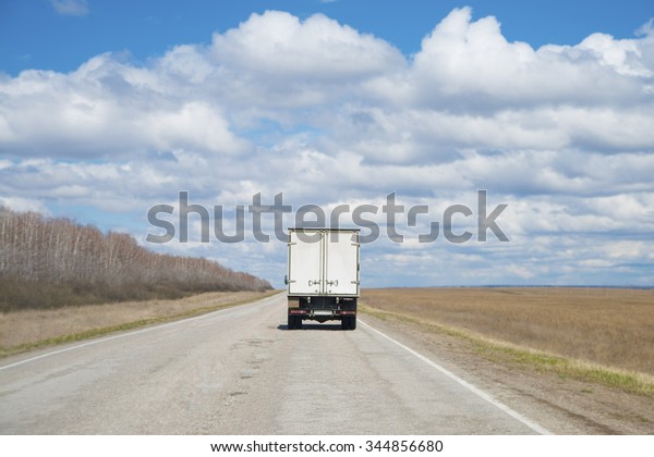 Alone\
Dirty Truck driving on clear spring asphalt road against blue sky\
with white clouds, empty field with dry grass and forest along\
highway Back view car Idea symbol of\
logistics