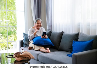 Alone Retired Asian Older Woman Sitting On Sofa And Talk To Her Family By Mobile Phone Is Using Internet At Living Room, Relax Time At Home.