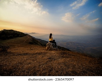 Alone tourist on the edge of cliff and watching into deep valley bellow at Phu Bak Dai, Loei, Thailand. Woman sit alone at the cliff of mountain to see scenic view of sunset and range of mountain. - Powered by Shutterstock