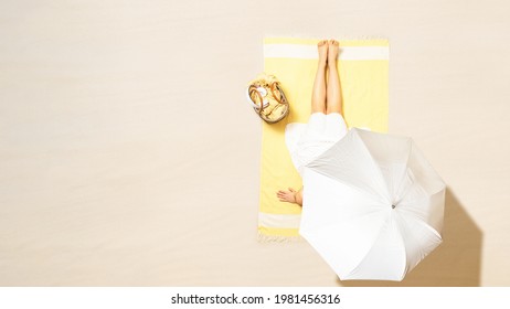 Alone Woman In Dress Sitting And Sunbathes On Yellow Beach Towel Under Umbrella With Bag. Female Relaxation On The Sand Beach At Summer Vacation. Top, Aerial View