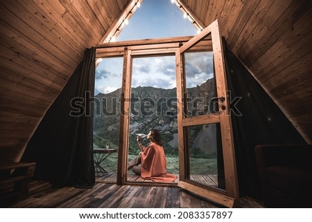 Alone woman drinking water on the porch of a small wooden house in the mountains. The concept of glamping and idyllic holidays