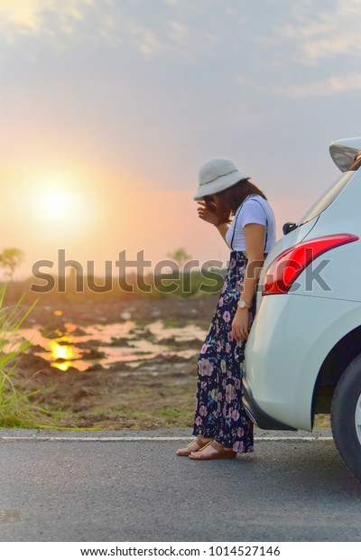 Alone Woman leaning back at\
hatchback car on road and feel sad against the sunset sky\
background.