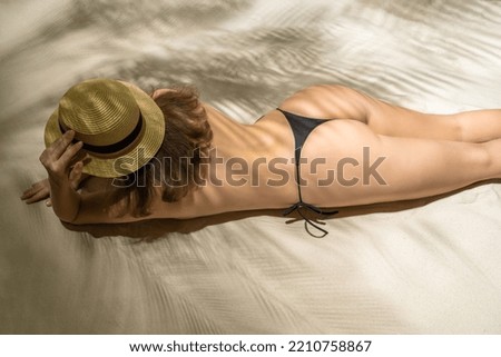 Alone woman in straw hat and bikini lying and sunbathes in a shadow under palm tree branches. Female relaxation on the sand beach at summer vacation. Top view