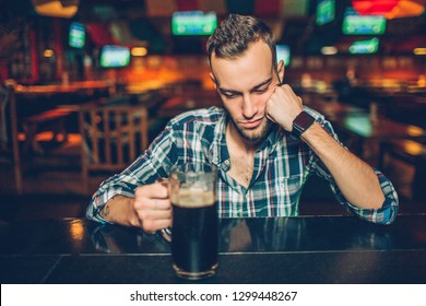 Alone Young Man Sit At Bar Counter In Pub. He Sleep. Young Man Hold Hand On Mug With Dark Beer.