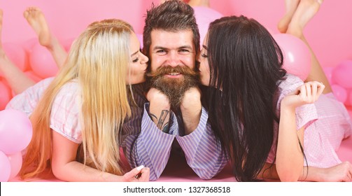 Alpha male concept. Threesome lay near balloons, happy guy on smiling face. Man with beard and mustache attracts blonde and brunette girls. Girls fall in love with macho, kissing, pink background.