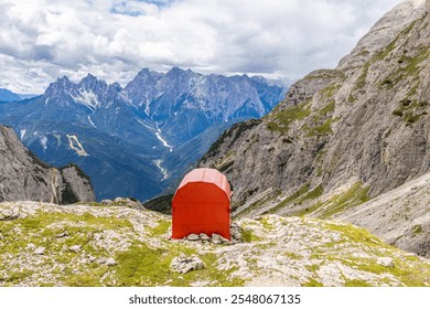 Alpine bivaco mountain hut shelter located in rugged alpine terrain. Red bivacco shelter in Dolomites. Compact structure against stunning landscapes, surrounded by peaks, valleys, and nature - Powered by Shutterstock