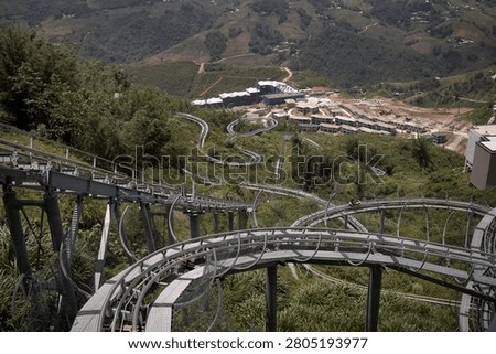 An Alpine Coaster track winds through a lush mountain landscape with twists, turns, and steep drops. This thrilling attraction is located on a hillside in Sa Pa, Vietnam, offering breathtaking panoramic views of the valley and surrounding hills. The metal track is built seamlessly into the natural terrain, showing how riders can coast downhill surrounded by green nature and distant buildings.