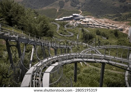 An Alpine Coaster track winds through a lush mountain landscape with twists, turns, and steep drops. This thrilling attraction is located on a hillside in Sa Pa, Vietnam, offering breathtaking panoramic views of the valley and surrounding hills. The metal track is built seamlessly into the natural terrain, showing how riders can coast downhill surrounded by green nature and distant buildings.