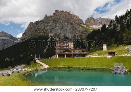 An alpine retreat hotel in the mountain by a turquoise lake with lush greenery and deep green pine trees, in front of a rugged mountain range 