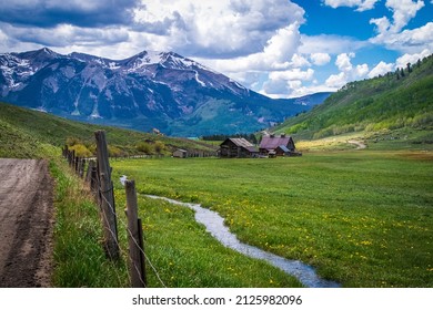 Alpine Scene With Dirt Road Rustic Barn And Snow Runoff Stream Running Through Valley With Wildflowers - Snow Peaked Mountains Near Crested Butte Colorado USA