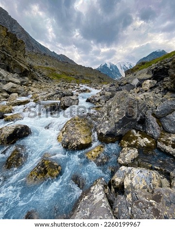 Alpine stream flows over stones near mountains with glaciers and snow in Altai in Siberia behind rainy clouds. Vertical frame.