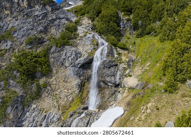Alpine waterfall cascading down rugged rocky cliffs into a mossy ridge, framed by evergreens in British Columbia, Canada.