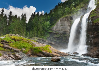 Alpine Waterfall In Mountain Forest Under Blue Sky.