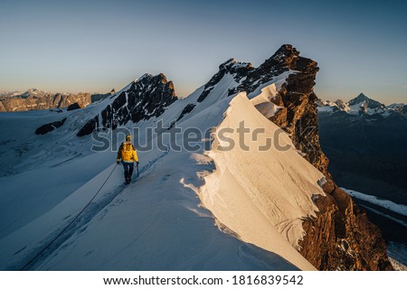 An alpinist climbing a rocky and snow mountain ridge during sunrise. Mountaineering and alpinism in Switzerland. Ascent of Breithorn, Zermatt. Alpine mountain landscape with snow and rocks.