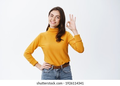 Alright. Smiling young woman hold all under control, show okay sign and looking confident, guarantee quality, advertising good product, standing against white background.
