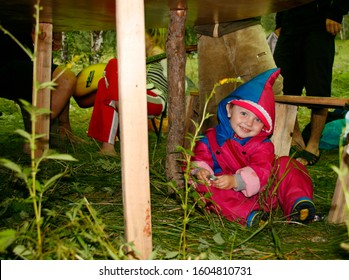 Altai, Russia - July 21, 2009. Parents With Young Children Go Camping In The Forest. A Small Child Sits On The Grass Under An Impromptu Table In Nature, Amid Adult Legs In The Forest. Love Of Nature.