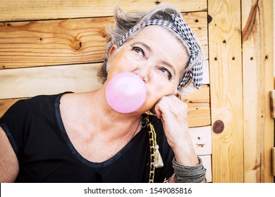 Alternative Unusual Pretty Senior Caucasian Lady With Trendy Make Up And Grey Hair Blowing A Pink Bubble Gum And Looking Up - Nice Portrait Of Old Youth People Enjoying The Life