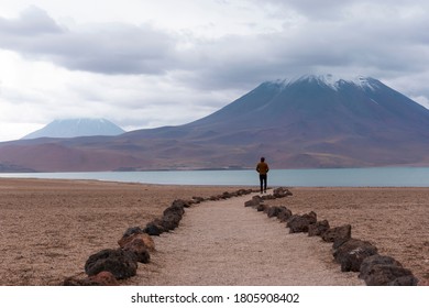 The Altiplano Of The Antofagasta Region, In Northern Chile. Woman Walking Outdoors With Lake And Volcano And Mountain View In The Background. Miñiques Volcano. Lagunas Miscanti Y Miniques.