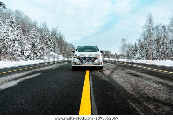 Aluksne, Latvia - 01.12.2021\
metallic white Nissan Leaf on asphalt road with yellow lines in\
winter. compact five-door hatchback electric car manufactured by\
Nissan