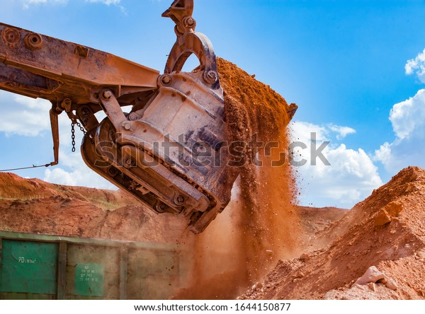 Aluminium ore quarry. Bauxite clay open-cut\
mining. Loading an ore to rail hopper car with excavator. Close-up\
of excavator bucket on blue sky with\
clouds.