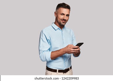 Always Available. Good Looking Young Man In Smart Casual Wear Using His Smart Phone And Smiling While Standing Against Grey Background