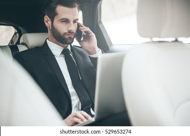 Always Available. Handsome Young Businessman Working On His Laptop And Talking On The Phone While Sitting In The Car