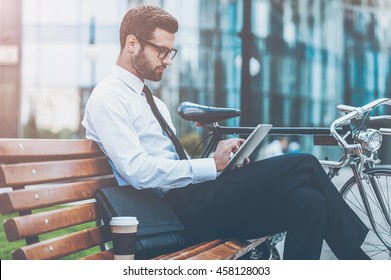 Always connected. Side view of confident young businessman working on digital tablet while sitting on the bench near his bicycle with office building in the background - Powered by Shutterstock