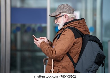 Always Connected. Side View Profile Of Pleasant Bearded Old Man In Glasses Is Standing With Backpack And Using Mobile Phone While Sending Message. He Is Looking At Screen With Smile