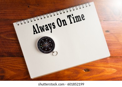 Always On Time words on notebook with compass over wooden background.