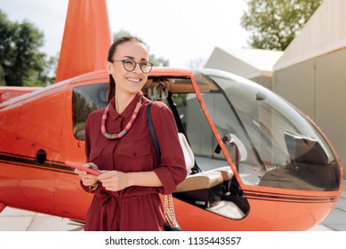 Always online. Low angle of merry optimistic woman posing near helicopter and using phone