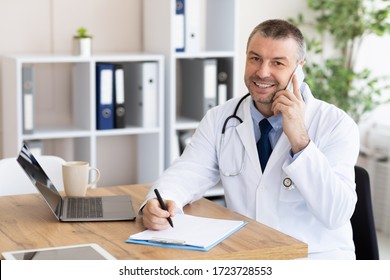 Always Ready To Help. Happy doctor talking on phone with his patients and writing notes, working at his office