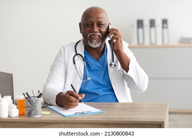 Always Ready To Help. Happy Mature African American Male Doctor Talking On Cell Phone With Patients And Writing Notes, Working At Clinic Office Sitting At Desk, Posing Looking At Camera